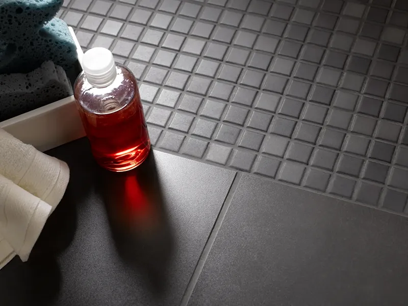 Dunkle Kleinmosaikfliesen grenzen an größere matte Fliesen, daneben liegt eine rote Flasche.